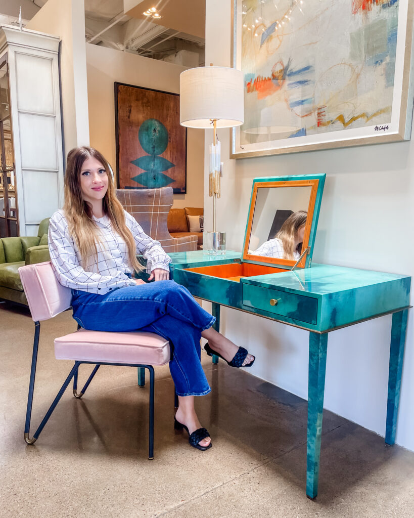 Individual sitting by a sleek, turquoise desk with a flip-up mirror in a stylishly decorated room, featuring modern artworks and elegant furniture.